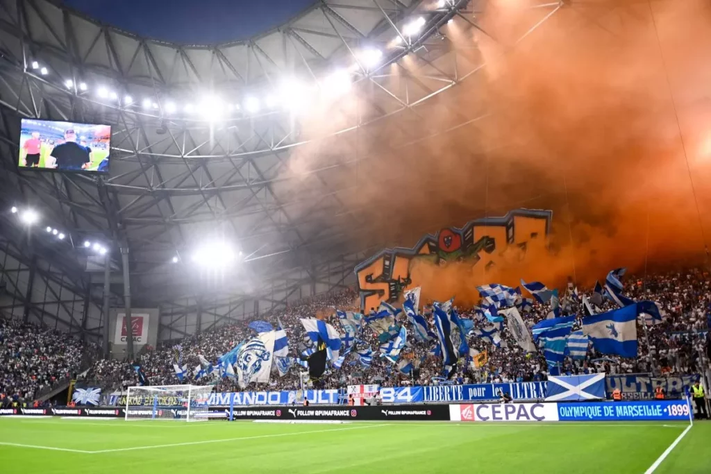 Image du Stade Vélodrome de l'Olympique de Marseille en plein match, avec les tribunes remplies de supporters en bleu, blanc et orange. Les drapeaux flottent au vent, et les lumières du stade illuminent l'atmosphère vibrante, symbolisant la passion des fans marseillais